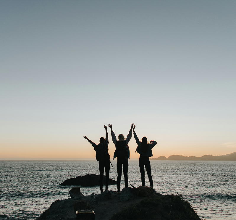 Silhouetten von drei Frauen, die bei Sonnenuntergang jubelnd mit erhobenen Armen auf einem Felsen am Meer stehen.