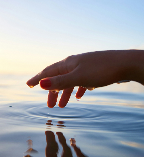 Eine Hand mit rot lackierten Fingernägeln berührt sanft die Wasseroberfläche eines Sees bei Sonnenuntergang, wodurch feine Wellen entstehen.